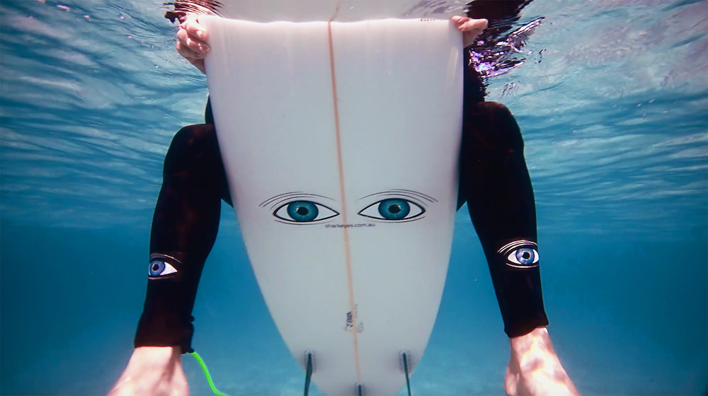 view of a surfer underwater sitting on surfboard half submerged in water with the Shark Eyes decal visible on bottom